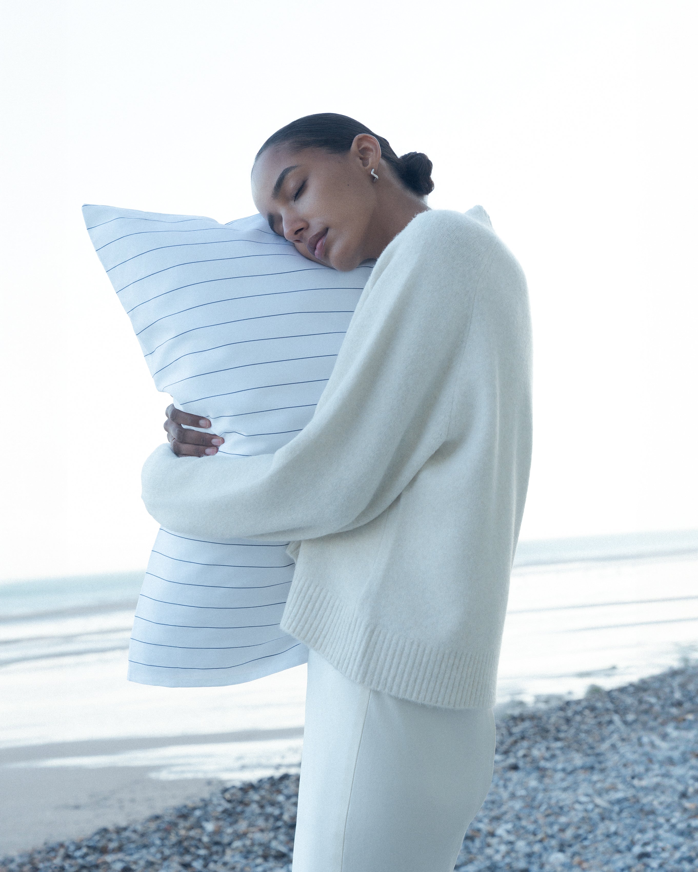 Female model with a hair bun and beige sweater, hugging a pinstriped soft pillow, standing on the beach.