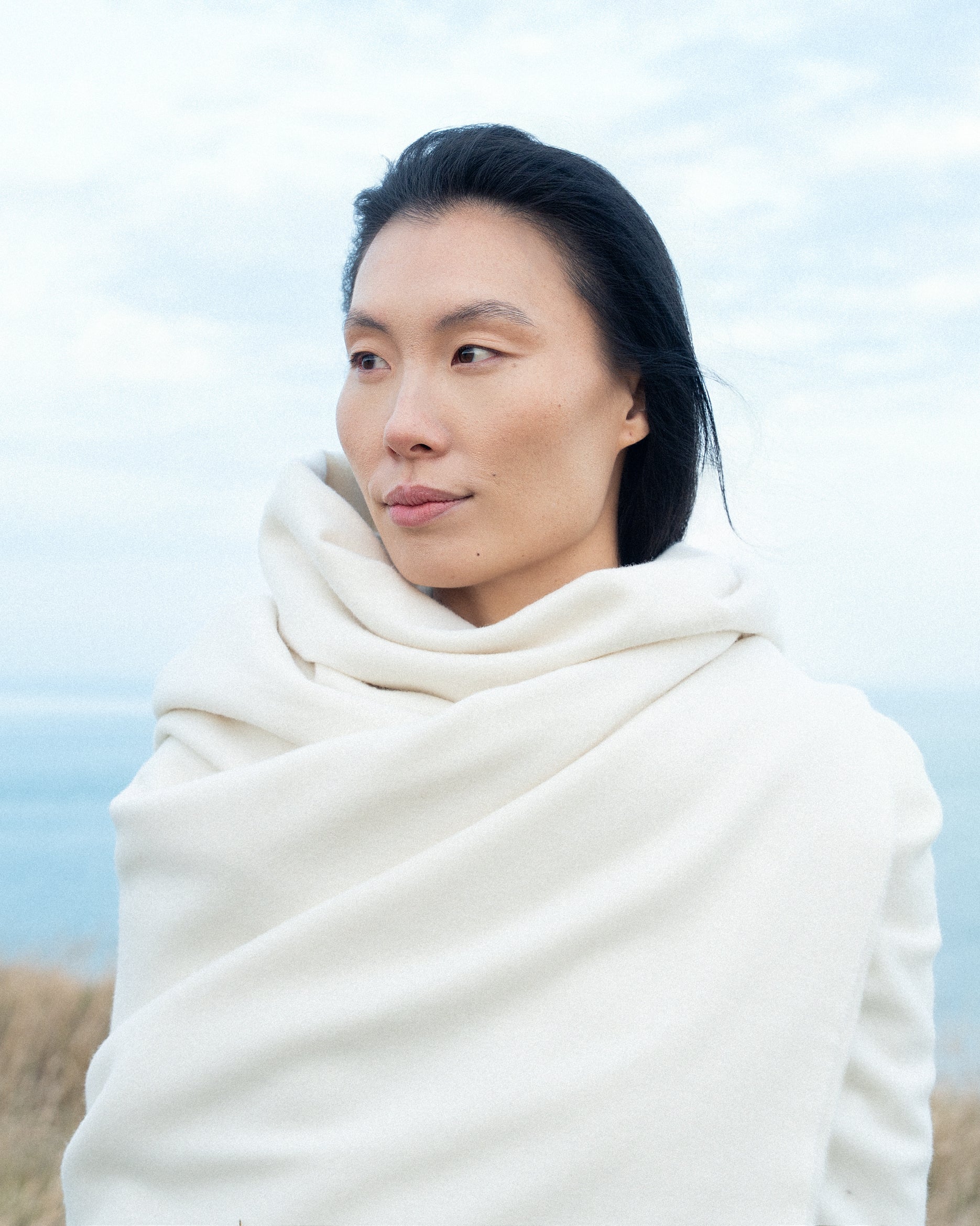 Dark haired female model looking away from the camera with a slight smile, wrapped around with a soft merino wool throw from Abate.
