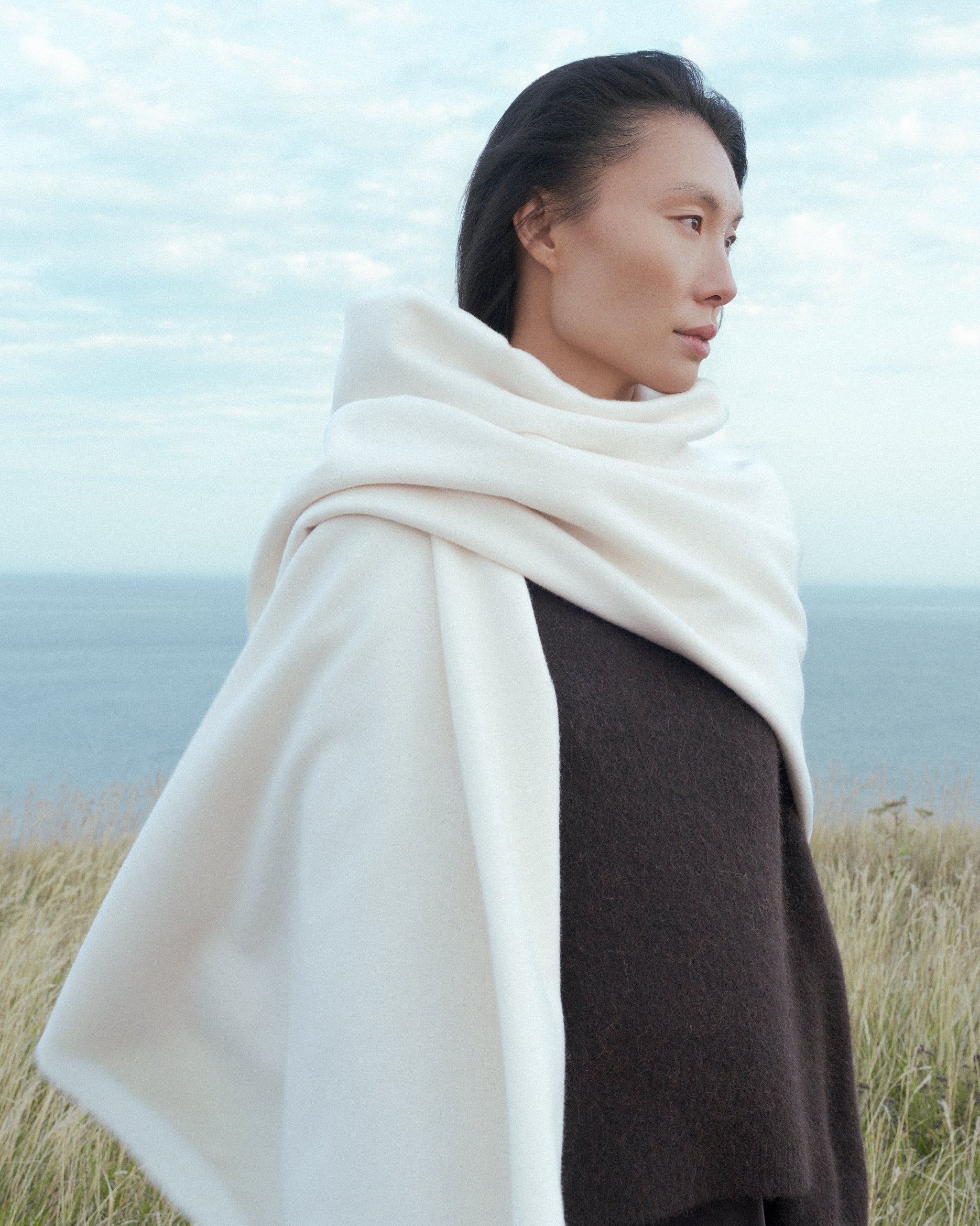 Dark haired female model looking on to a field with a ocean background, wrapped around with a soft merino wool throw from Abate.