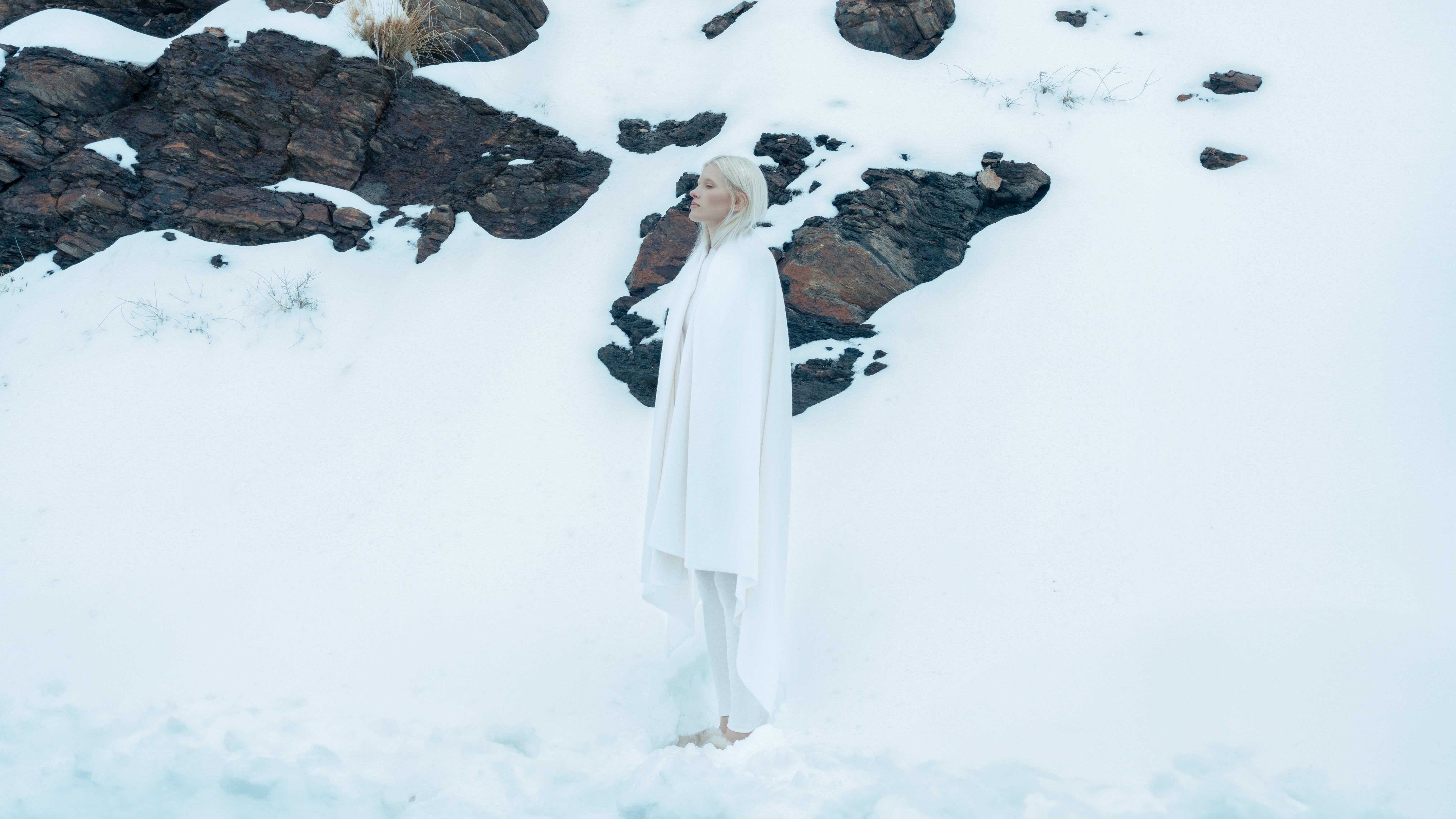Woman standing in showy landscape with white hair and white eyebrows, standing in a showy landscape with a white merino wool throw draped around her shoulders.