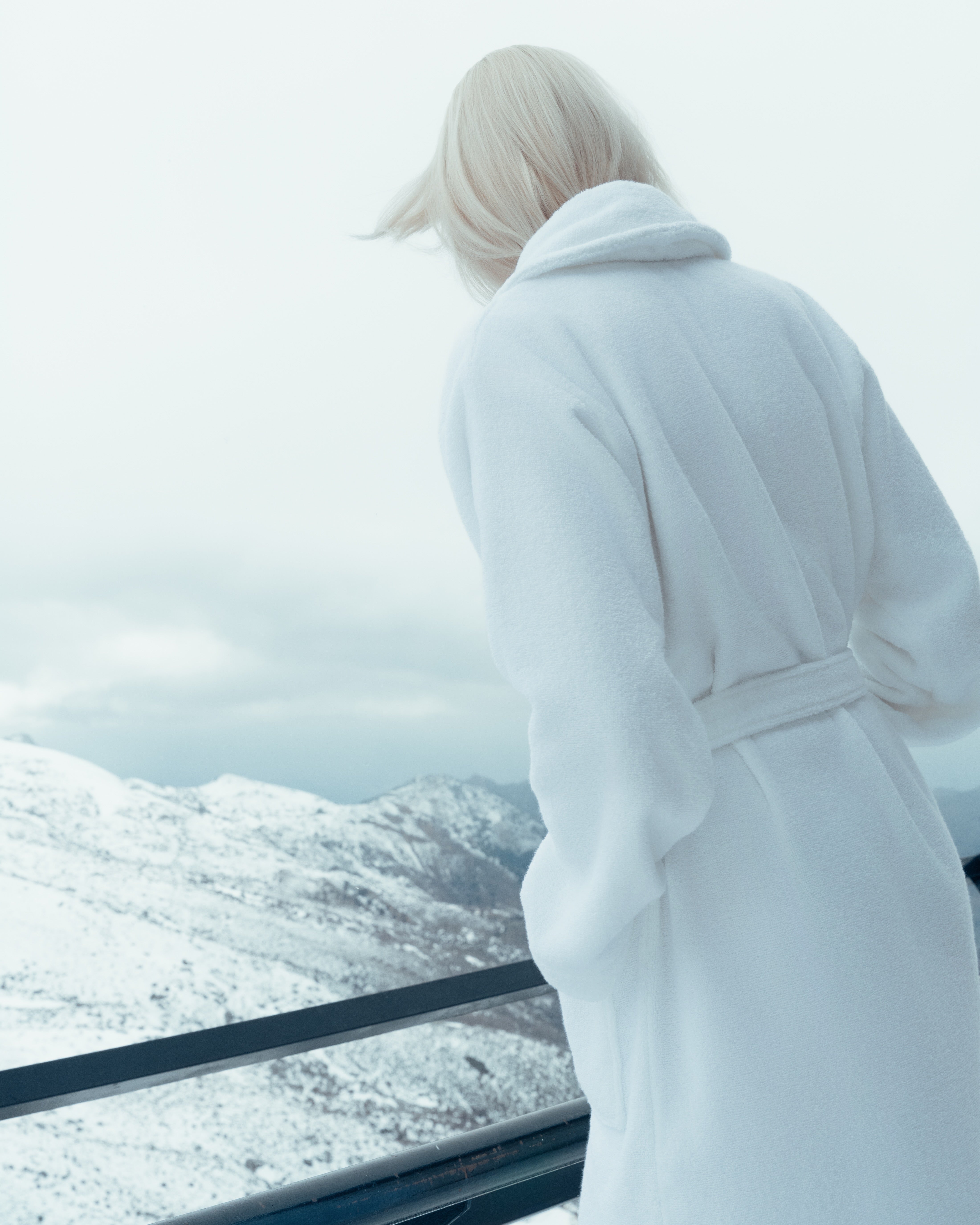 Woman leaning over a guardrail, looking down at a winter landscape with blue skies, wearing a white terry bath rope.