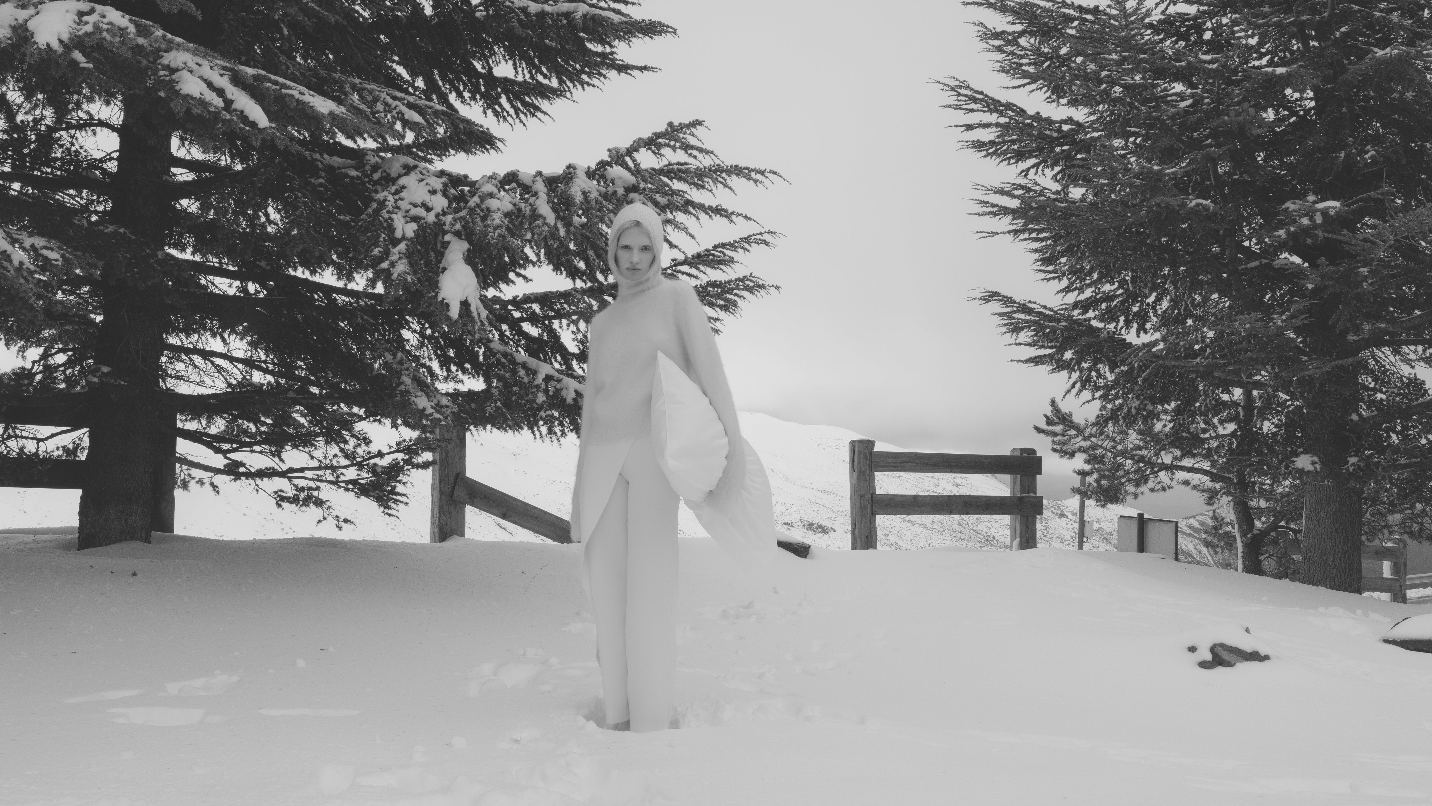 Black and white image of a woman, standing in a snowy landscape with trees behind her, dressed in a light outfit and head scarf, holding a soft pillow case in her left arm.