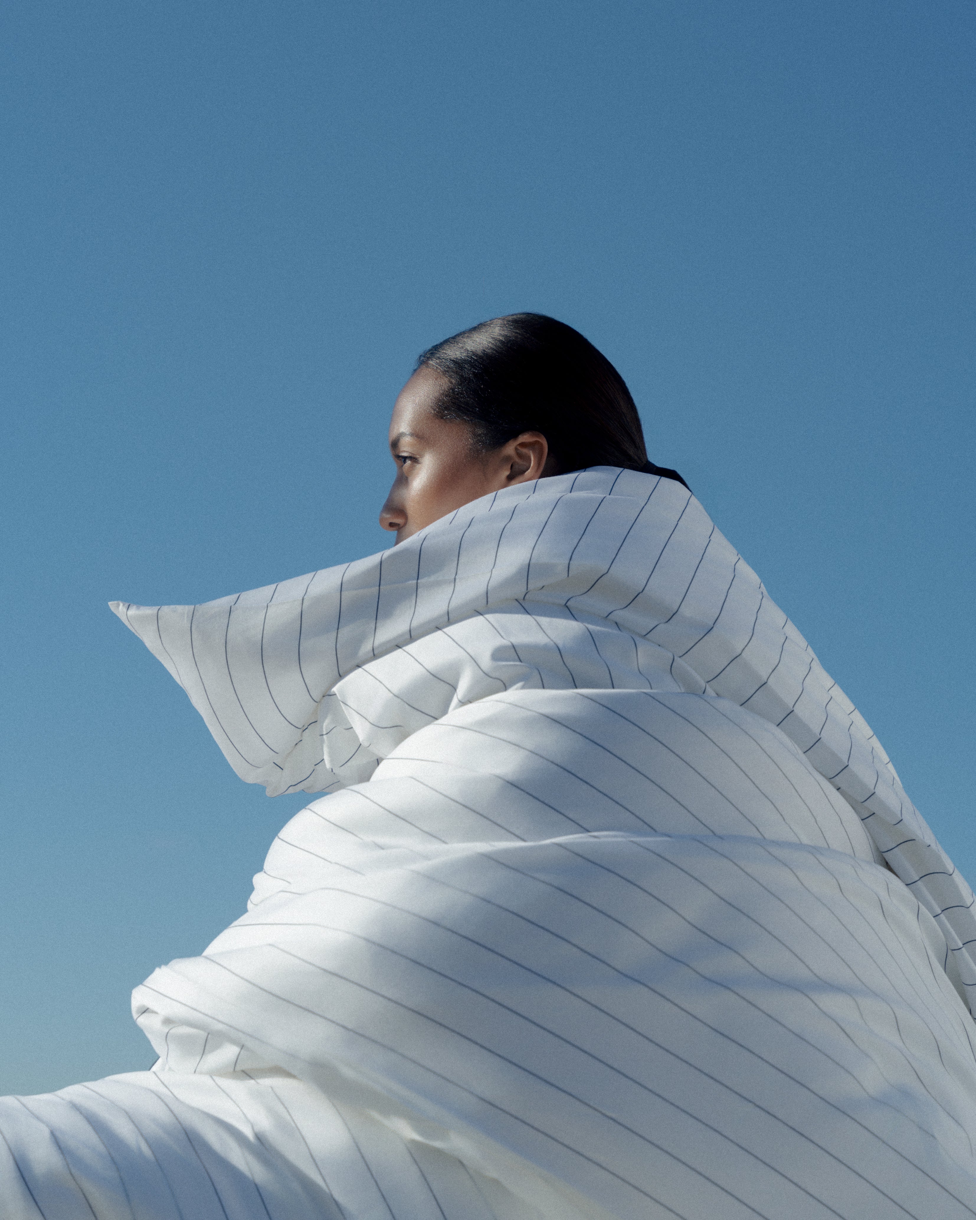 Image with model wrapped in a huge striped duvet with a blue background and soft sun on her face