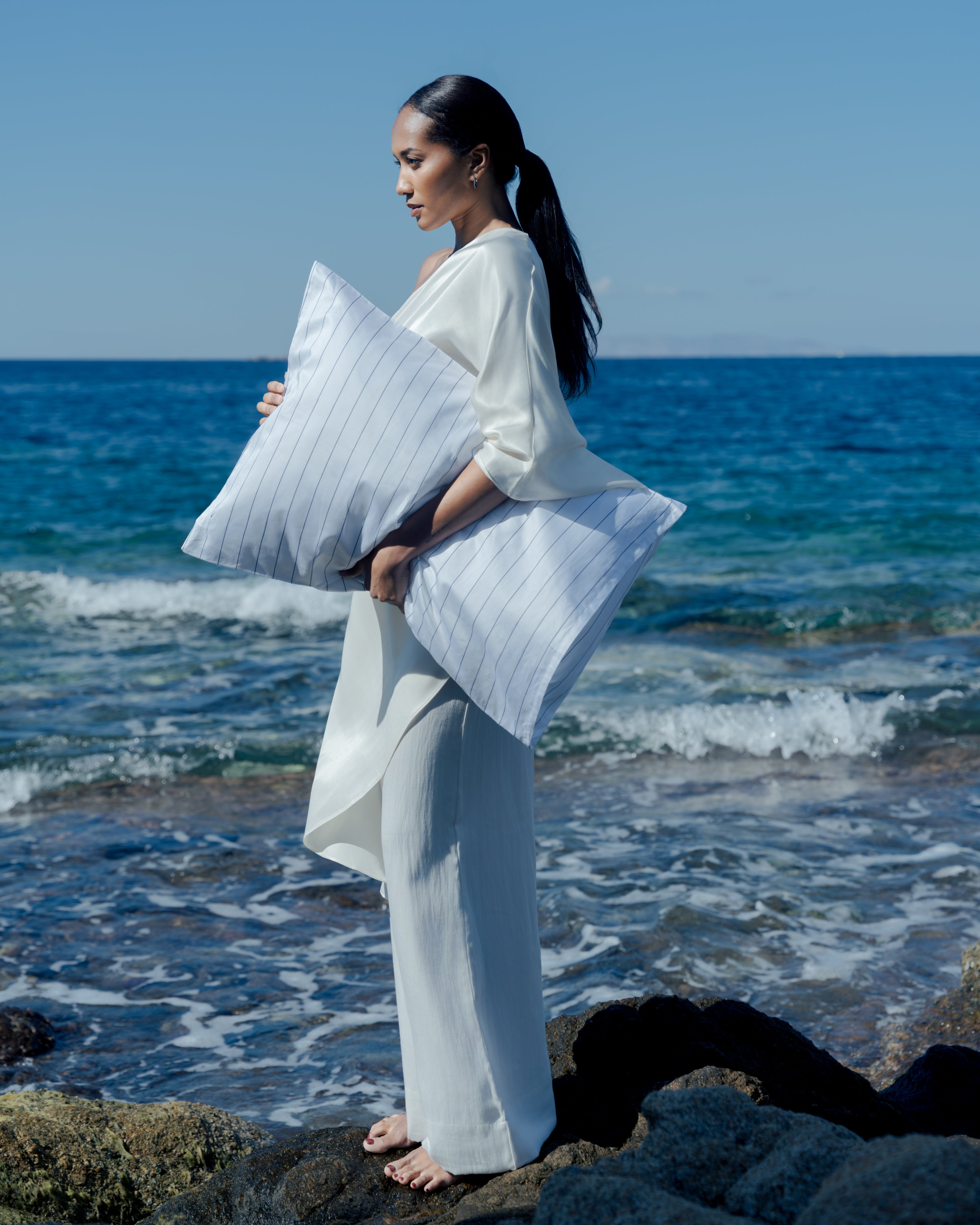 woman standing in front of the ocean, on a sunny day, holding a blue striped pillow.