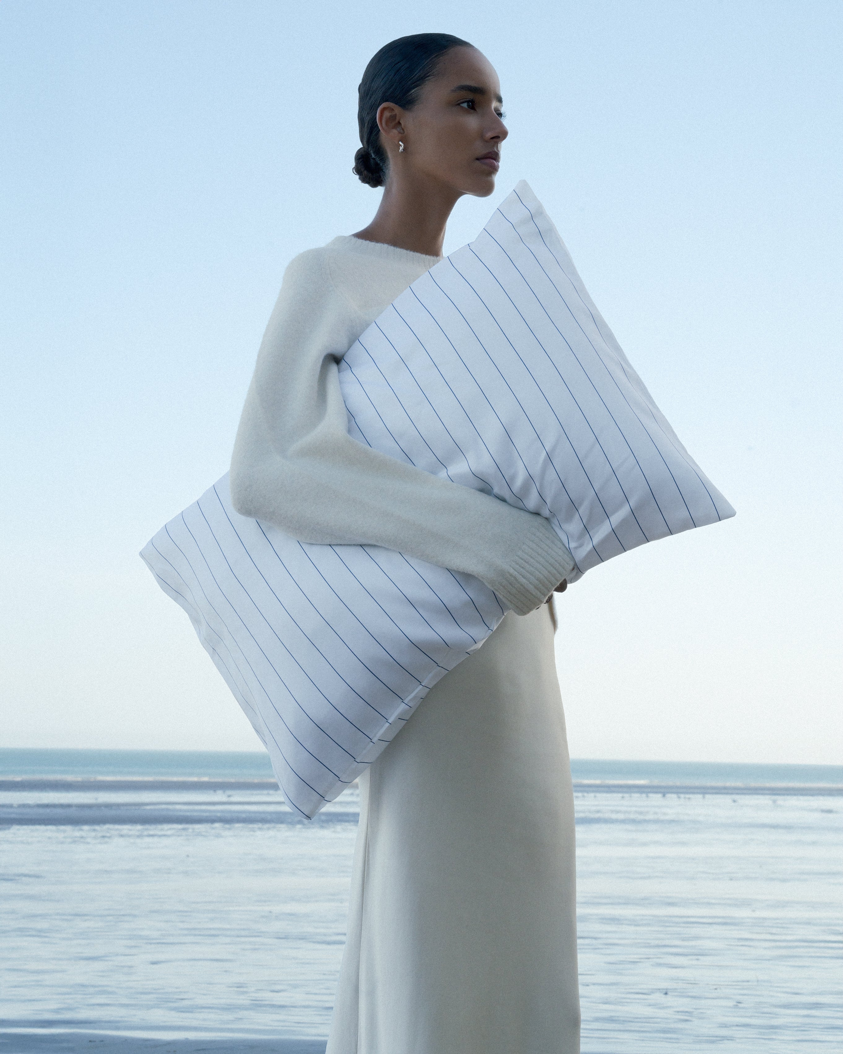 Dark haired woman, in a light beige skirt and sweater, holding a striped soft pillow with the ocean and blue skies behind her.