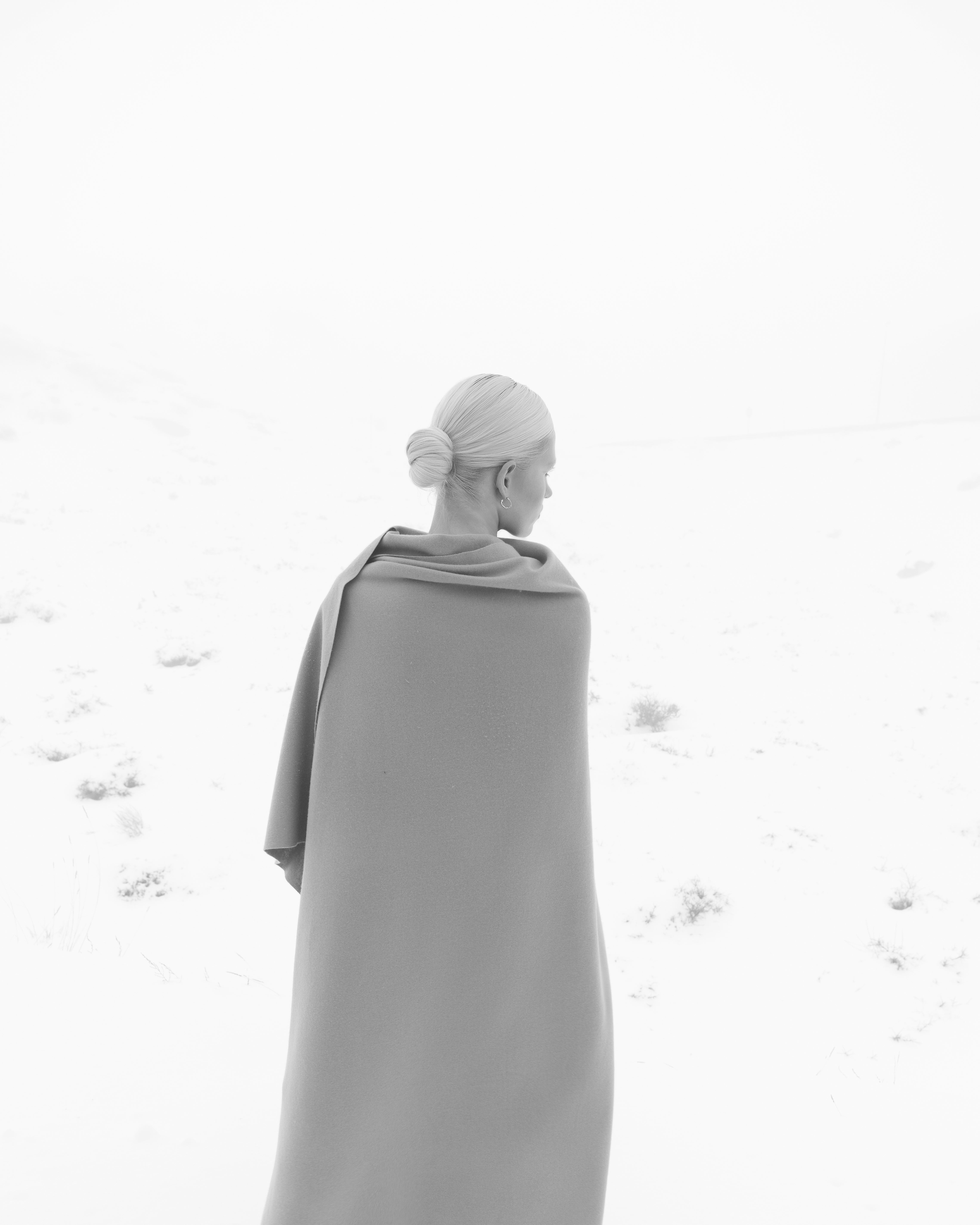 Woman standing with her back to the camera, in a white snowy landscape and a merino wool throw covering her shoulders and body. 