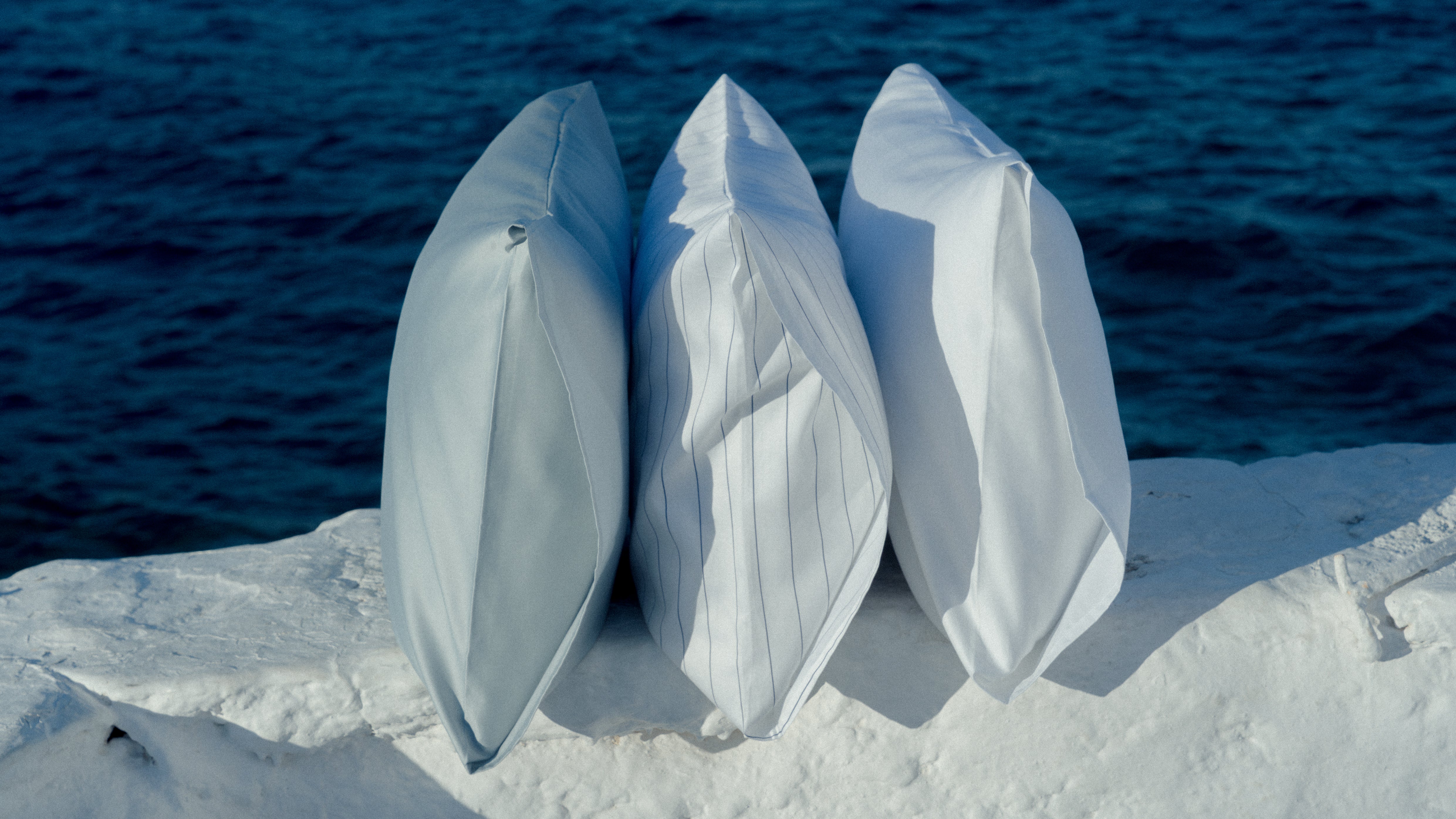 3 pillows standing next to each other in light colors with the blue ocean behind them.